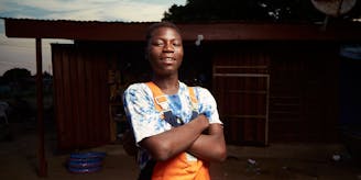 Portret van Teye Abrantie (18). Hij staat trots met zijn armen over elkaar in zijn monteurskleding, voor de monteursschool. Hij lacht in de camera.